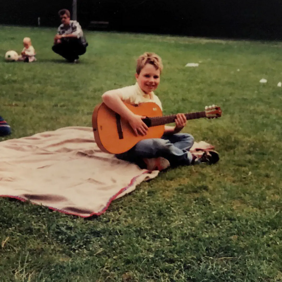 Me as a kiddo, smiling happily with my new guitar, sitting on a picnic blanket in the park