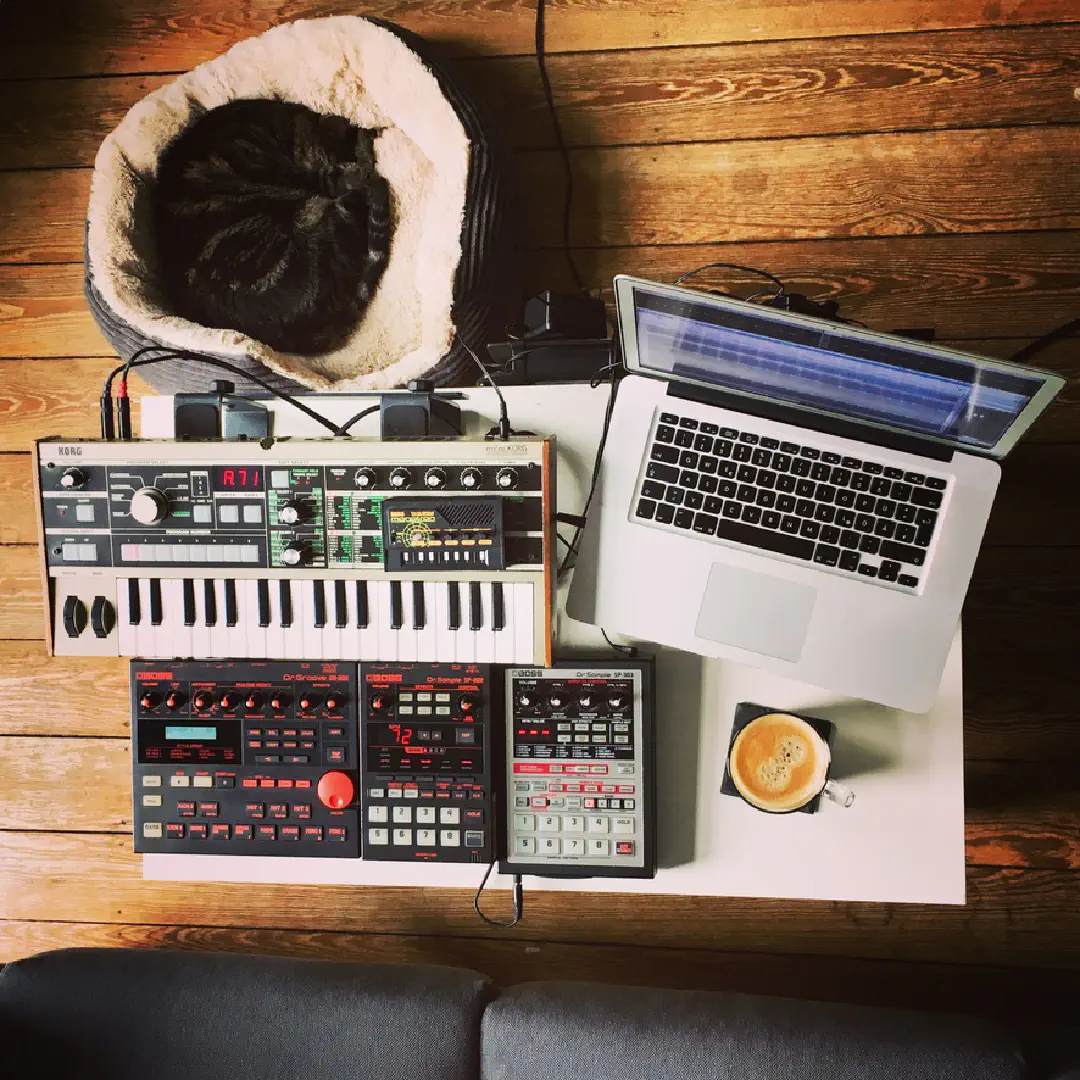 Top view of a coffee table, filled with a synthesizer, a drum computer, two samplers, and a macbook. A cat is sleeping next to it.